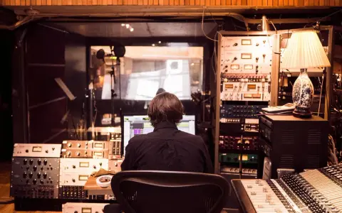 Osoba siedzi przed komputerem w profesjonalnym **recording studio**, otoczona sprzętem audio i mikserem.