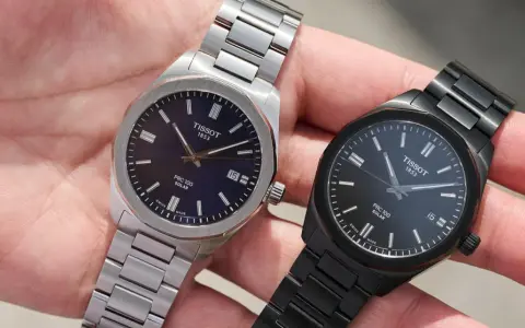 Dwa zegarki Tissot PRC 100 Solar na dłoni. Jeden srebrny z granatową tarczą, drugi czarny z czarną tarczą. Oba posiadają mechanizm solarny.
