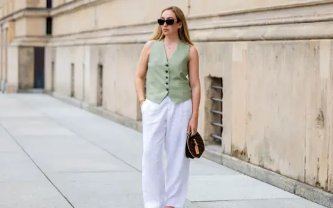 Mujer con un outfit de chaleco verde y outfit pantalon lino blanco, gafas de sol y bolso de mano.