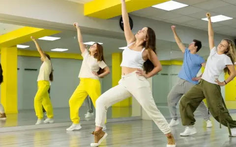 Grupa młodych ludzi ćwiczy taniec w sali z lustrami. Ich ruchy oddają energię gatunku muzycznego pop.