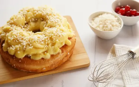 Rosca de Pascua decorada con crema pastelera y azúcar granulada, lista para disfrutar. A su lado, cuencos con cerezas y ralladura, y un batidor.