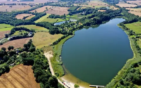 Malownicze jezioro Marszewo otoczone zielenią i polami. Widać piaszczystą plażę z pomostem i kilka samochodów.