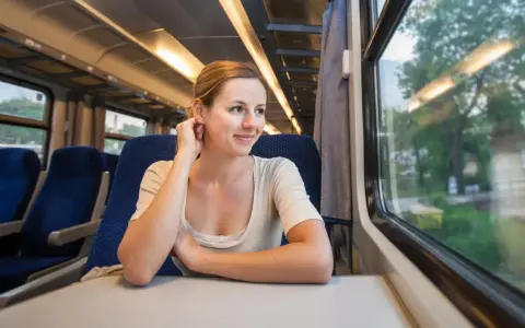 Uśmiechnięta kobieta podróżuje pociągiem, podziwiając krajobrazy, które mogą być częścią najpiękniejszych tras kolejowych w Europie.