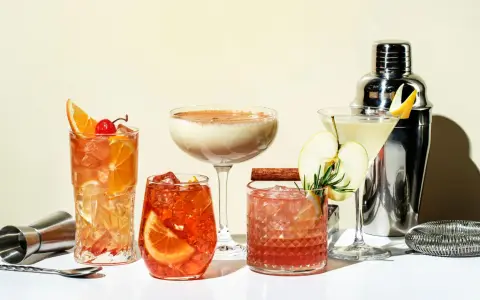 Eine Auswahl an fruchtigen Cocktails, garniert mit Orangen, Äpfeln und Kirschen, neben einem Cocktail-Shaker.