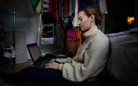 Ung kvinna sitter på golvet och jobbar på sin laptop, hon kombinerar studera på distans och jobba samtidigt.