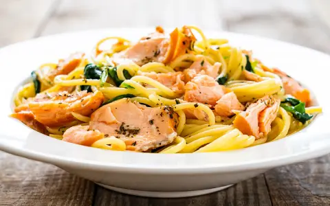Deliciosa pasta con salmón desmenuzado y espinacas, servida en un plato blanco sobre una mesa de madera.