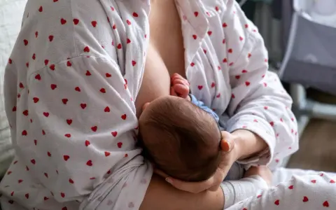 Matka karmi piersią dziecko. W tle widać łóżeczko. Zatkany kanał mleczny to częsty problem.
