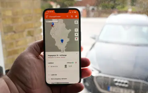 En hand håller upp en mobil med appen ChargeFinder som visar en karta över laddstationer för elbil. En laddapp visar en laddare nära en elbil.