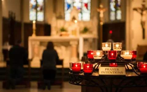 Candles glow in a church, with a sign reading "Prayer Intentions." People kneel in prayer, their mass intentions illuminated by the soft light.
