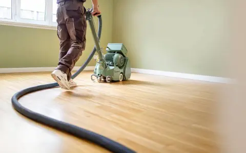 Mann bearbeitet einen Holzboden mit einer Schleifmaschine. Der Parkettboden schleifen ist eine wichtige Arbeit für die Renovierung.