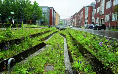 Grüne Oase mit blauer Infrastruktur: Ein Stadtgarten mit Wegen aus Kopfsteinpflaster und metallischen Beeteinfassungen, umgeben von üppiger Vegetation und Blumen.
