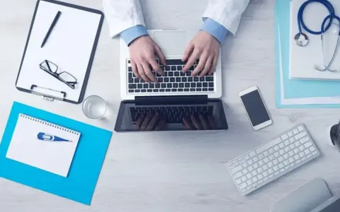 Lekarz pisze na laptopie, obok stetoskop, recepta, telefon i termometr.