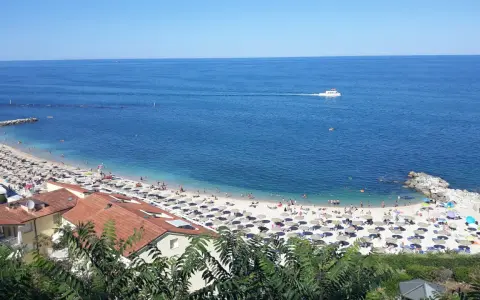 Le spiagge di Numana, con ombrelloni colorati e gente che si gode il sole, bagnate da un mare azzurro e calmo. Una barca solca l'orizzonte.