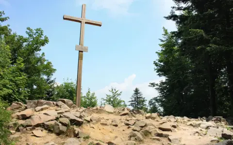 Drewniany krzyż na skalistym szczycie, najwyższy szczyt w górach świętokrzyskich, otoczony zielenią drzew.