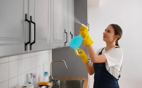 Kobieta w żółtych rękawiczkach czyści meble z okleiny, używając sprayu i ściereczki.