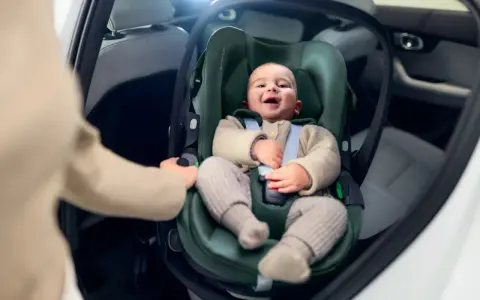 Uśmiechnięte dziecko w foteliku samochodowym. Ręka rodzica pokazuje, jak wypiąć fotelik z bazy isofix.