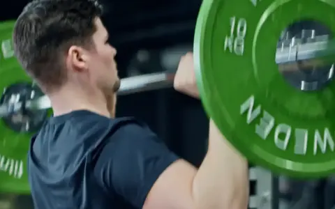 Man utför axelpress med skivstång. Gröna viktskivor med texten "SWEDEN" och "10 KG" syns tydligt.