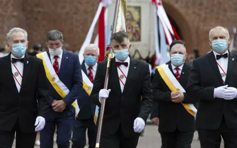 Rycerze Kolumba w garniturach i maskach maszerują z flagą.