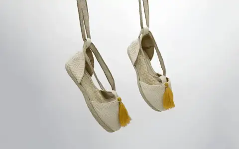 Dos alpargatas de tela beige con pompones amarillos cuelgan de tiras, mostrando su diseño veraniego.