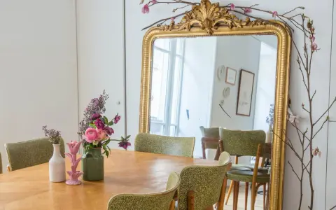 Un grand miroir ancien doré, orné de branches fleuries, reflète une salle à manger avec des chaises vertes. Quelle couleur peindre un miroir ancien ? L'or reste un classique intemporel.