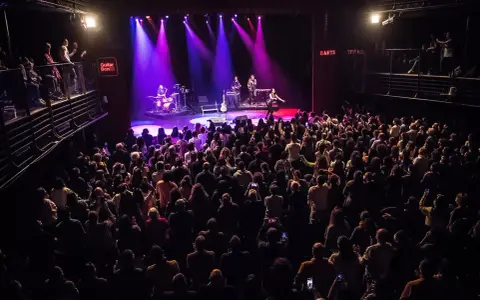 Un concierto en Guitar BCN ilumina el escenario con luces púrpuras mientras una multitud entusiasta graba el momento.