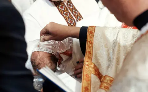 Ksiądz w bogato zdobionej szacie chrzci dziecko, polewając mu głowę wodą z kryształowej karafki.