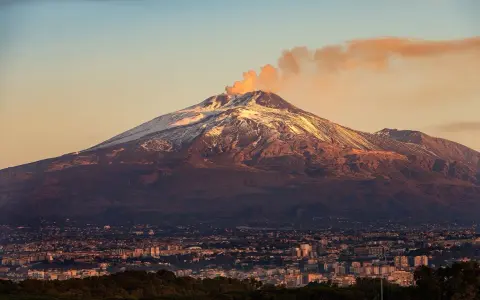 Etna fumante, con la cima innevata che brilla al tramonto, domina una città siciliana.