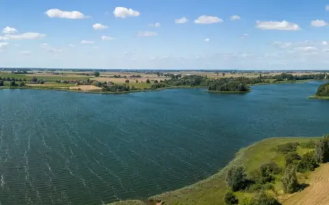 Malownicze jezioro Lednica z wyspą pośrodku, otoczone zielenią i polami. Błękitne niebo z białymi chmurami.