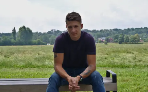 Bartłomiej Bis siedzi na ławce w parku, w tle zielone drzewa i niebo.