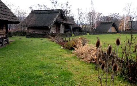 Park archeologiczny Osada VI Oraczy. Drewniane chaty z krytymi strzechą dachami, otoczone plecionymi płotkami, na tle jesiennej zieleni.