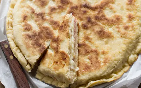 Focaccia in padella dorata e fragrante, tagliata a spicchi per mostrare il ripieno filante. Un coltello con manico in legno è appoggiato accanto.