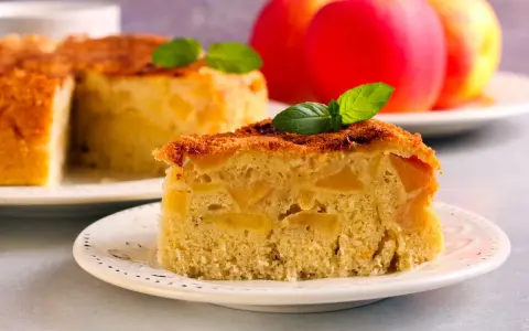 Una fetta di torta di mele vegana, soffice e ricca di mele, guarnita con menta. Sullo sfondo, una torta intera e mele rosse.