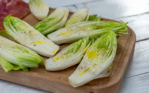 Insalata belga tagliata a metà, condita con olio e pepe, su un tagliere di legno.