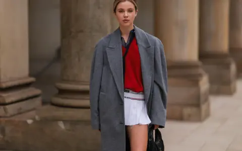Ein junges Model präsentiert einen preppy look mit grauem Mantel, rotem Pullover und weißem Faltenrock.