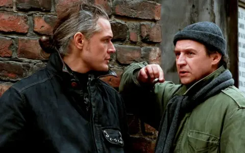 Dwóch mężczyzn rozmawia przy ceglanej ścianie. Jeden z nich, z siwymi włosami związanymi w kok, pali papierosa. Czy to rozmowa o tym, czym jest ekstradycja?