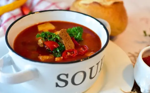 Aromatyczna zupa gulaszowa w białej misce z napisem "SOUP", ozdobiona natką pietruszki i papryką. Idealna na chłodne dni, to jedne z tych **dobrych zup domowych**.