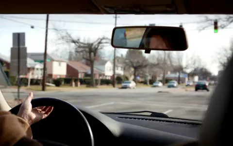 Mano sul volante, pronta per una manovra di svolta. Riflesso del volto nel retrovisore, case e alberi spogli fuori dal finestrino.