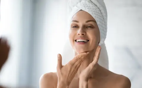 Une femme souriante, serviette sur la tête, applique délicatement de l'eau micellaire sur son visage pour une peau fraîche et nette.