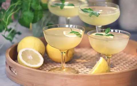 Fyra glas med en ljusgul limoncello drink, garnerade med mynta och citron. Citroner ligger på en träbricka.