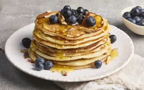Stapel fluffiger High Protein Pancakes mit Blaubeeren und Mandelsplittern, beträufelt mit Honig.
