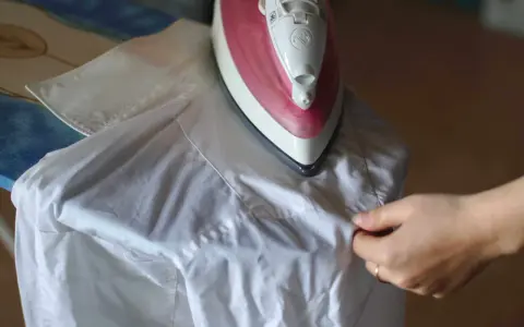 Różowe żelazko na desce do prasowania, obok biała koszula. Dłoń poprawia materiał, pokazując, jak prasować koszule.
