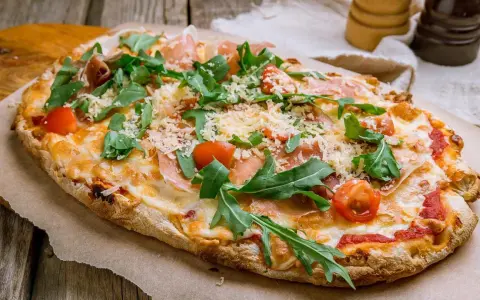 Eine köstliche Pinsa belegt mit Schinken, Rucola, Kirschtomaten und viel Käse auf rustikalem Holzuntergrund.