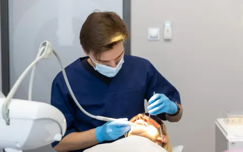 Dentysta w niebieskim fartuchu i masce wykonuje zabieg. Pacjentka ma założone okulary ochronne. Nowoczesne znieczulenie komputerowe zapewnia komfort.