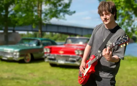 Erik Meduza spelar gitarr framför klassiska bilar vid en sjö.