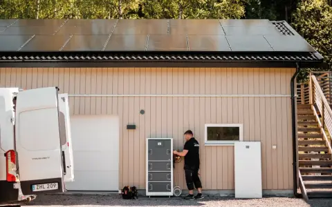En tekniker installerar ett batterilager för dt energi vid ett hus med solpaneler på taket.