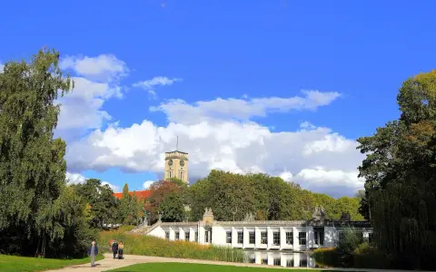 Ein sonniger Tag im **Rudolph Wilde Park**. Ein langer, weißer Pavillon spiegelt sich im Wasser, während ein Uhrenturm über Bäumen aufragt.
