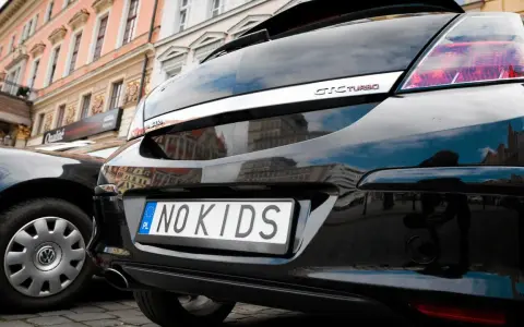 Czarne auto z indywidualnymi tablicami rejestracyjnymi "NO KIDS" na tle kamienicy.
