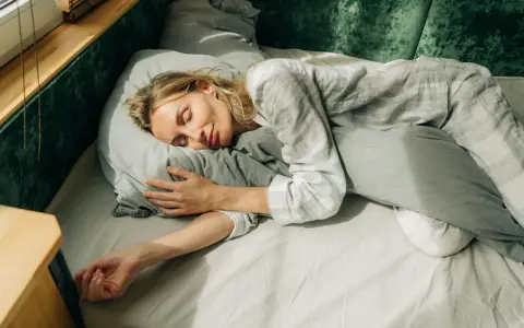 En kvinna sover lugnt i sängen, omfamnande en kudde. Solstrålar faller in genom fönstret och skapar en känsla av guidad avslappning.