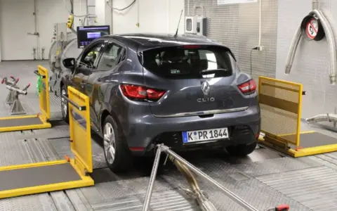 A dark grey Renault Clio is on a dynamometer in a testing facility, with exhaust pipes connected.