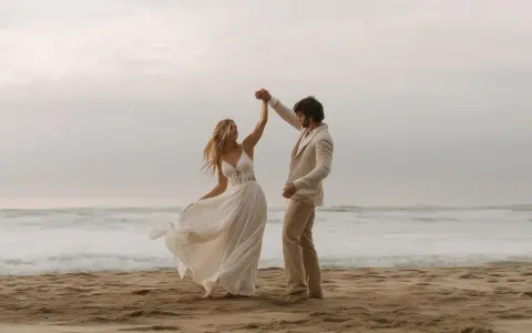 Coppia felice balla sulla spiaggia. Lui indossa un completo chiaro, perfetto per un matrimonio in spiaggia come vestirsi uomo.